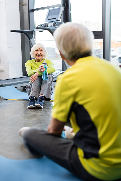 Smiling senior woman holding sports bottle near blurred man in gym