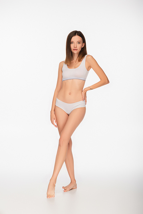 full length view of young and slim woman in lingerie standing with hand on hip on white background