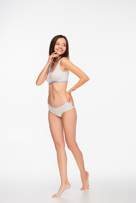 happy woman in underwear looking away while standing with hand on hip on white background