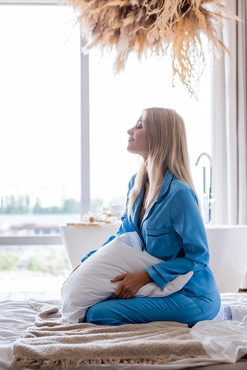 side view of joyful young woman holding pillow while sitting on bed