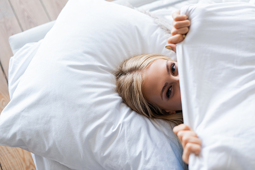 top view of young woman covering face with blanket while lying on bed