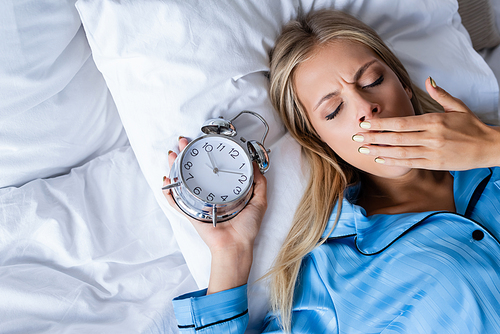 top view of woman covering mouth and yawning while lying with alarm clock in bed