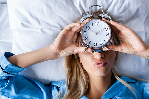 top view of blonde woman holding retro alarm clock and pouting lips while lying on bed