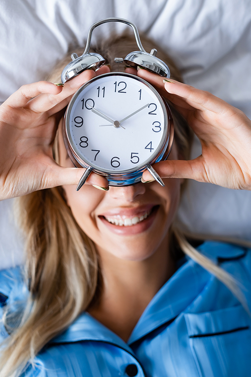 top view of cheerful woman holding retro alarm clock and covering face while lying on bed