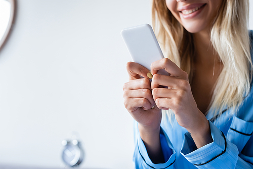 cropped view of cheerful woman in pajamas texting on smartphone in bedroom