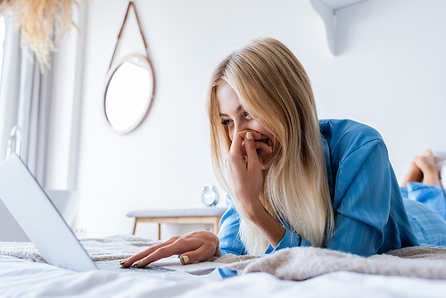 happy blonde woman in pajamas using laptop in bedroom