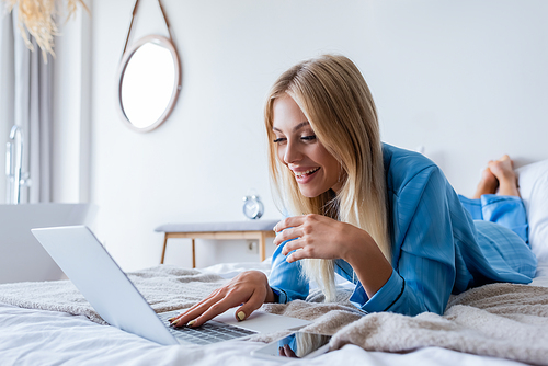 happy young freelancer in pajamas using laptop in bedroom