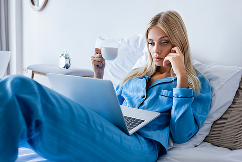 young woman in pajamas using laptop and holding cup of coffee