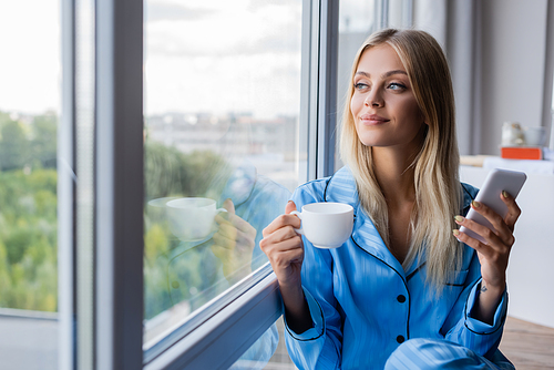 happy young woman holding cellphone and cup of coffee near window