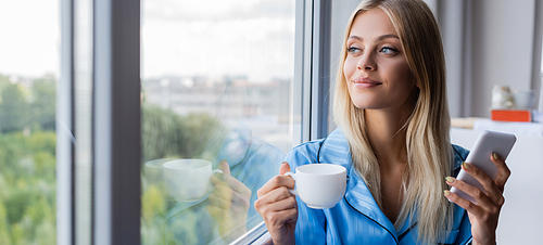 happy young woman holding cellphone and cup of coffee near window, banner