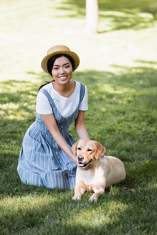 cheerful asian woman in striped sundress  near dog on lawn in park