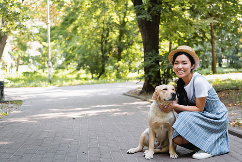 cheerful asian woman in straw hat  while hugging labrador in park