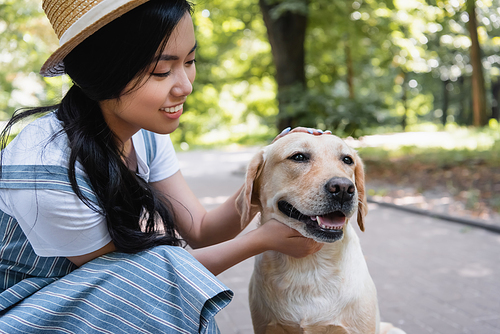 young asian woman smiling while stroking yellow labrador in park
