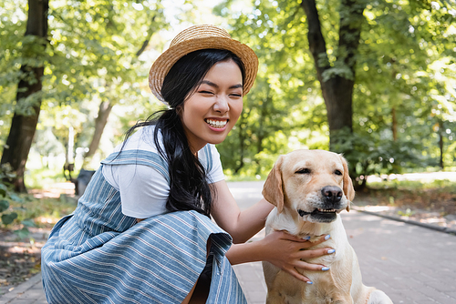 happy asian woman in straw hat embracing yellow labrador in park