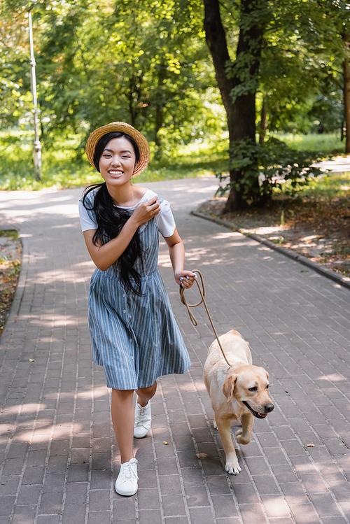 excited asian woman smiling at camera while strolling with dog in park