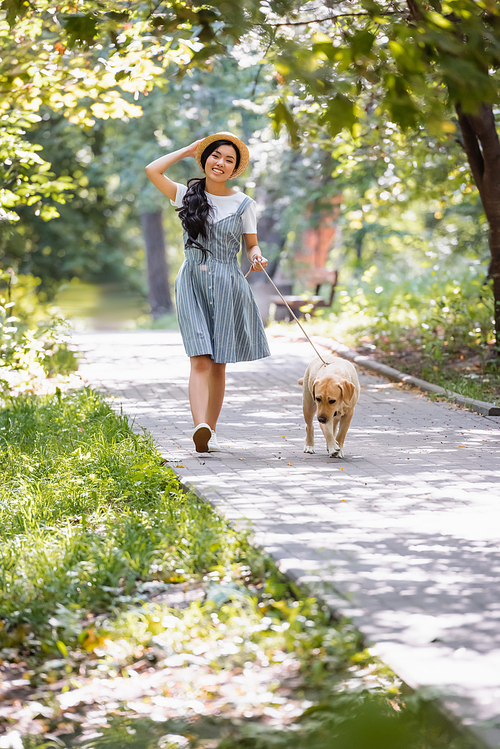 cheerful asian woman adjusting straw hat while walking with yellow labrador