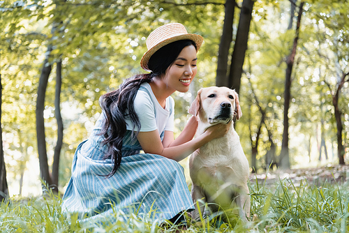 young asian woman in straw hat cuddling yellow labrador in park