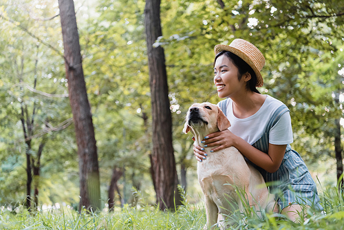 pleased asian woman in straw hat hugging dog and looking away in park