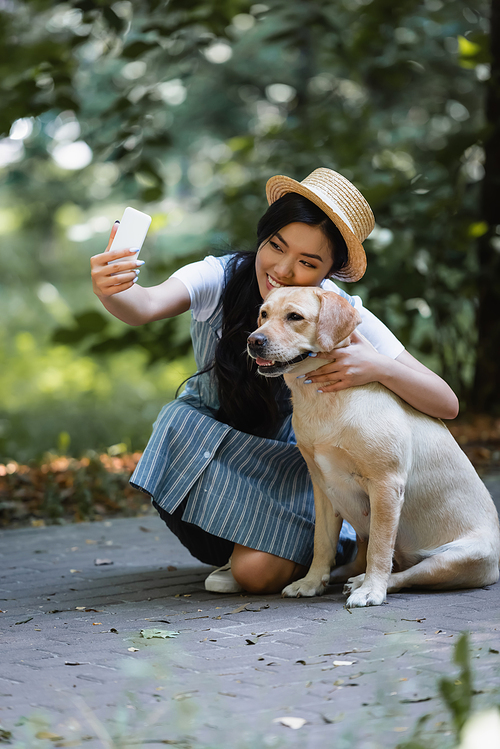 cheerful asian woman taking picture on smartphone with labrador in park