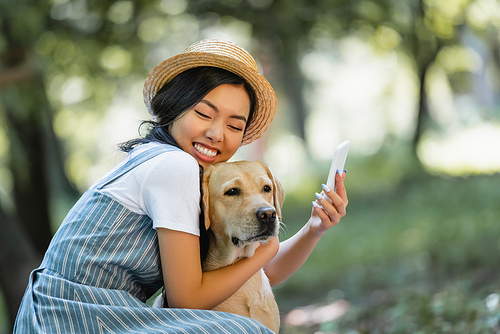 happy asian woman with closed eyes hugging labrador while taking selfie in park