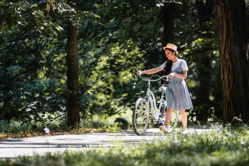 happy asian woman in striped sundress walking with bicycle in park