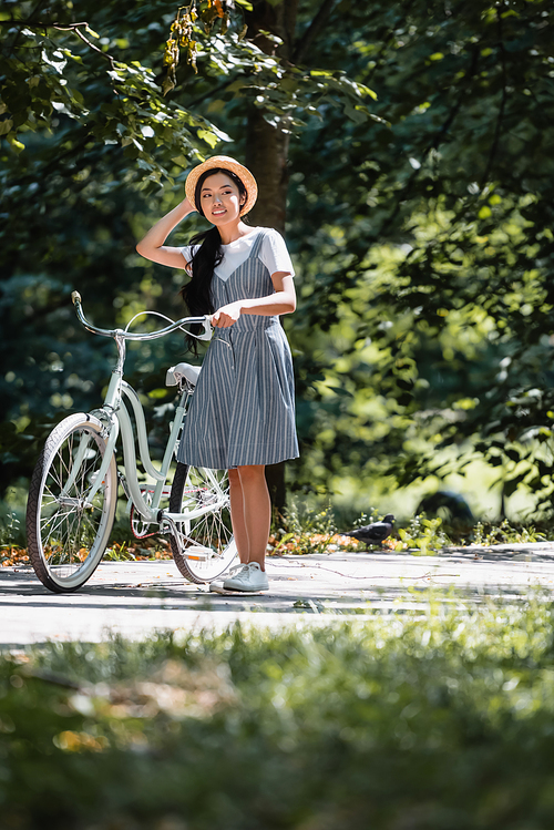 asian woman touching straw hat while looking away near bike in park