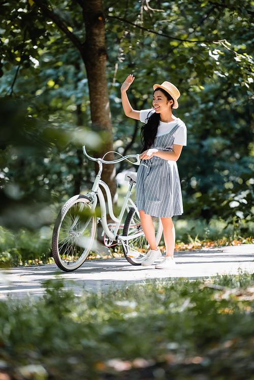 pleased asian woman looking away and waving hand near bike in park