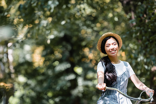 happy asian woman in straw hat smiling at camera while biking in park