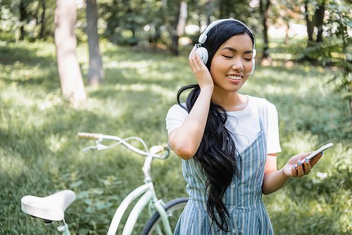 smiling asian woman adjusting headphones while using cellphone near blurred bicycle