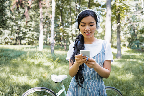 happy asian woman in headphones using mobile phone near blurred bike