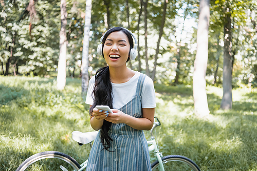excited asian woman with smartphone laughing in headphones near blurred bike
