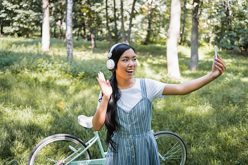 joyful asian woman in headphones waving hand while taking selfie near blurred bike