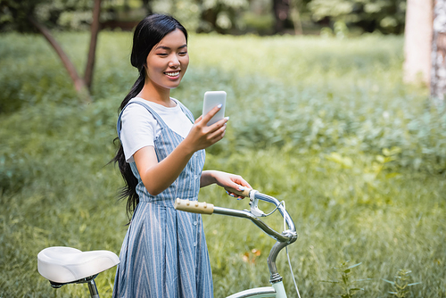happy asian woman in striped sundress looking at mobile phone near bicycle