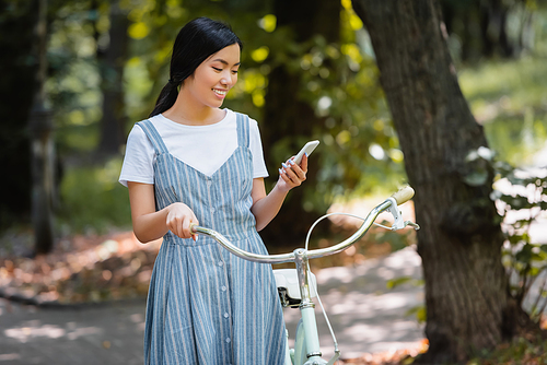 pleased asian woman with bicycle messaging on smartphone in park