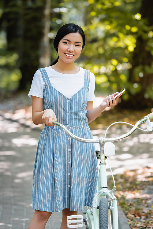 young asian woman with smartphone smiling at camera near bike in park
