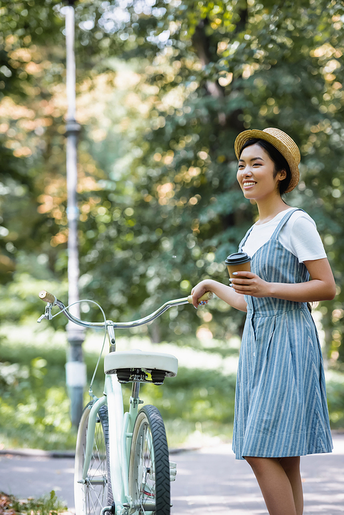 asian woman with paper cup looking away and smiling near bike in park