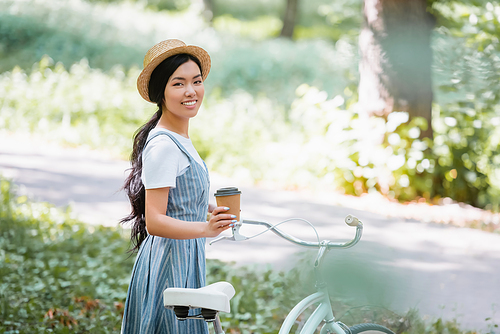 young asian woman in straw hat holding coffee to go near bicycle in park