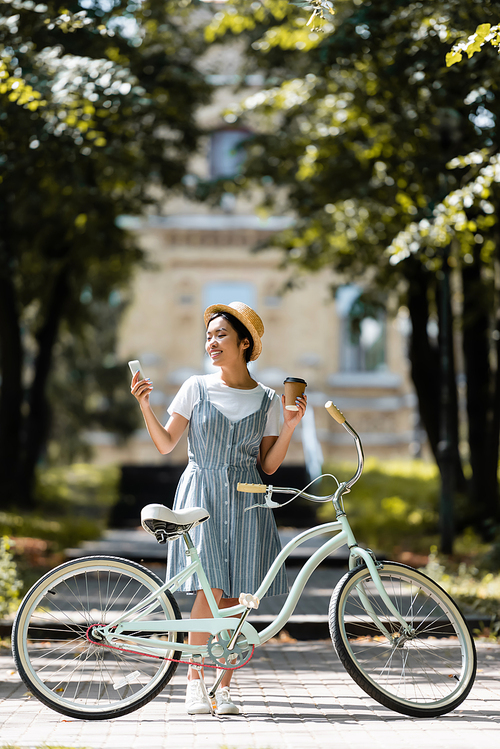 smiling asian woman with takeaway drink using smartphone near bike in park