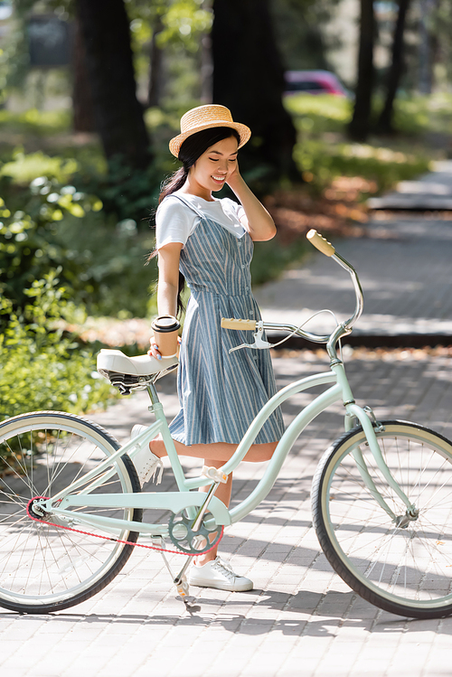 pleased asian woman in striped sundress standing near bicycle in park