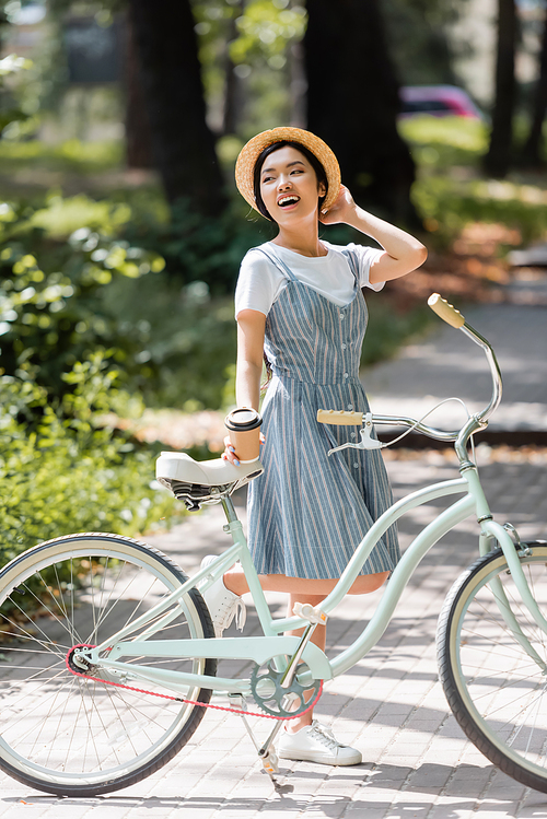 laughing asian woman touching straw hat while looking away near bike in park