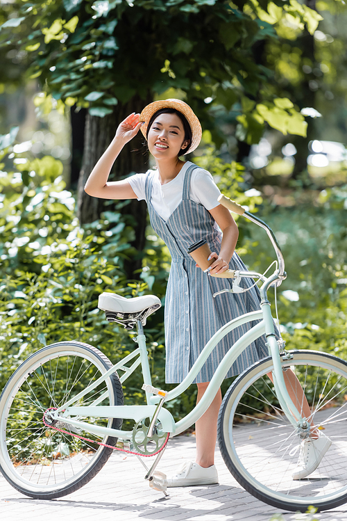 young asian woman adjusting straw hat while smiling at camera near bike in park