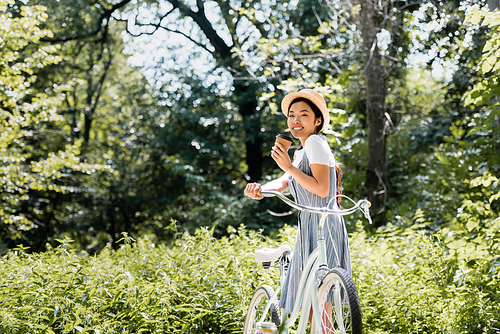 happy asian woman with bicycle and coffee to go smiling at camera in park