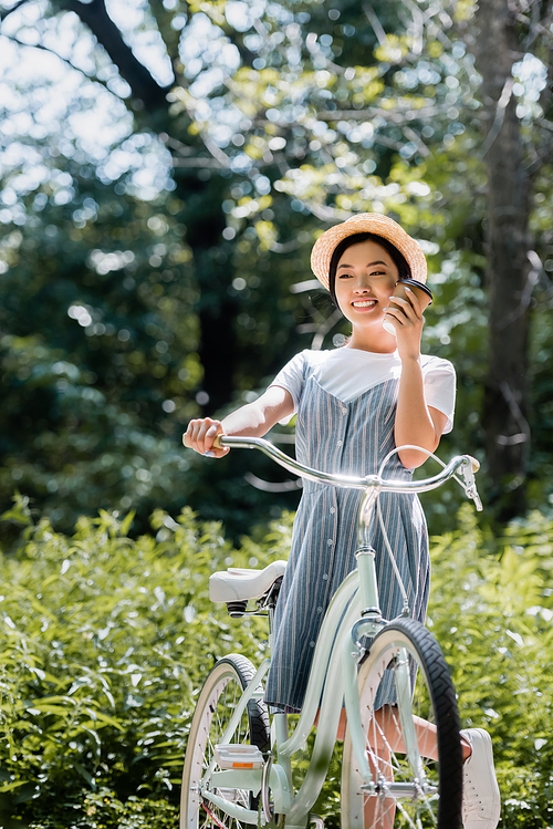 happy asian woman holding paper cup near face while standing with bike in park