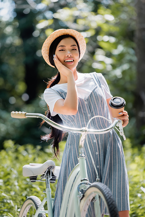 cheerful asian woman with coffee to go smiling at camera near bicycle in park