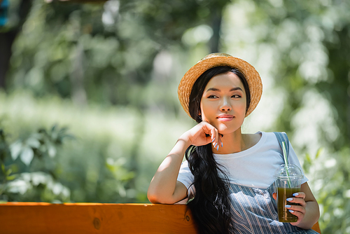 dreamy and smiling asian woman in straw hat sitting with fresh smoothie in park