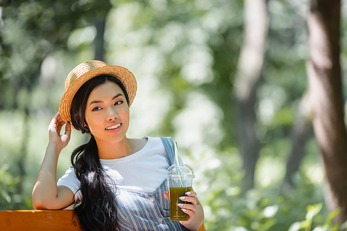 asian woman with fresh smoothie smiling while looking away in park