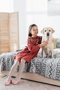 happy preteen girl cuddling labrador dog while sitting on bed at home