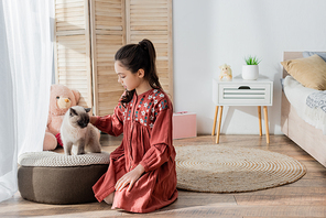 girl sitting on floor and stroking cat on soft pouf in bedroom