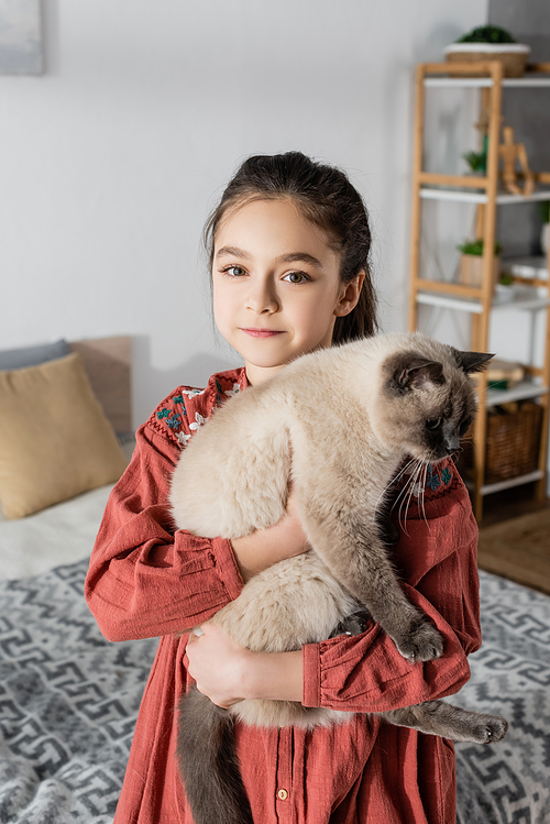 preteen girl smiling at camera while hugging cat at home