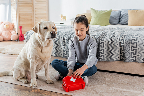 smiling girl playing doctor with toy first aid kit and labrador dog on floor in bedroom